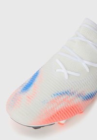 Fußballschuh mit einem weißen strukturierten Obermaterial, blauen und pinken Verlaufstreifen, weißen Schnürsenkeln und einer geformten Sohle mit mehreren Stollen.