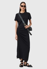 Abito t-shirt nero con maniche corte, dettaglio arricciato laterale e silhouette lunga. Abbinato a sandali neri flat e una borsa a cintura nera.