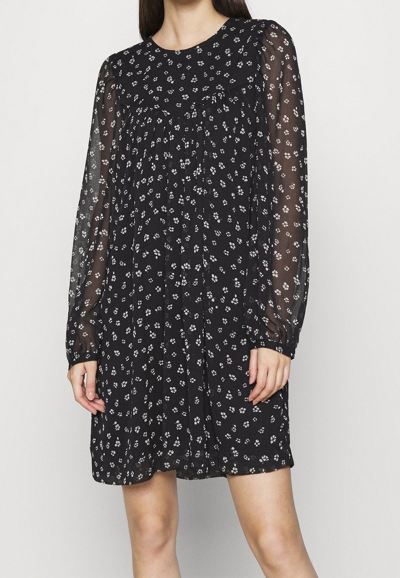 Femme portant une robe noire à manches longues transparentes avec un motif floral blanc, debout devant un fond clair uni.