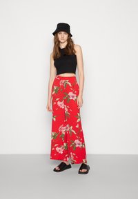 Top curto preto, calças largas vermelhas com flores rosa e verdes, acompanhadas de sandálias pretas. Chapéu simples estilo bucket.