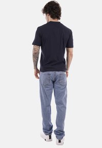 Uomo con capelli ricci e braccio tatuato che indossa una maglietta nera, jeans azzurri e scarpe da ginnastica bianche, visto da dietro.