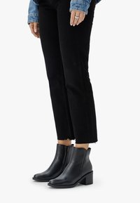 Eine Person trägt schwarze Stiefeletten mit Absatz, schwarze verkürzte Jeans mit fransigen Säumen und eine Jeansjacke, während sie vor einem weißen Hintergrund steht.