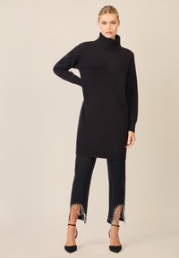 Schwarzer Rollkragenpullover mit langen Ärmeln und geripptem Saum. Kombiniert mit schwarzen Jeans, die an den Bündchen mit Perlenfransen verziert sind, und schwarzen Absatzschuhen.