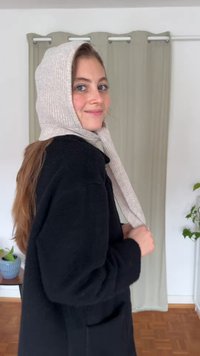 Bufanda de punto acanalado gris claro usada sobre un abrigo oscuro, cayendo hacia abajo, con una textura suave. El fondo presenta cortinas claras y una planta en maceta.