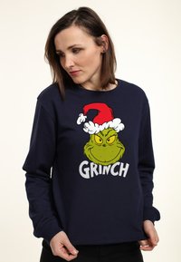 Sudadera azul marino con un gran gráfico del Grinch que lleva un gorro de Santa rojo y blanco, terminada con letras blancas en negrita debajo.
