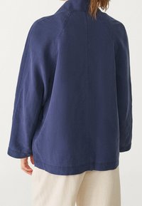 Donkerblauwe jas met een ontspannen pasvorm, lange mouwen en een detail met een naad aan de achterkant. Gemaakt van zachte stof, met een eenvoudig en strak ontwerp.