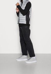 Graue, pufferartige Weste mit schwarzen Akzenten, getragen über einem schwarzen Langarmshirt. Das Outfit umfasst schwarze Hosen und graue Sneakers mit weißen Sohlen.