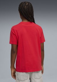 Άτομο με κοτσίδες, που φοράει απλό κόκκινο κοντομάνικο πουκάμισο και ανοιχτό γκρι τζιν παντελόνι, με την πλάτη προς τα εμπρός σε γκρι φόντο.