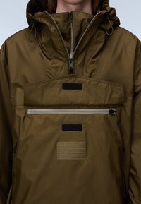 Person iført en olivengrøn hættetrøje med lynlåslomme foran, Velcro-mærker og et syet flagemblem på brystet.