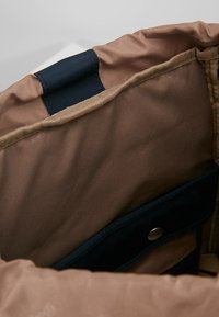 Wnętrze brązowej torby materiałowej z beżową podszewką, granatowymi akcentami oraz zapięciem na zatrzask. Zawiera boczną kieszeń o gładkiej teksturze.