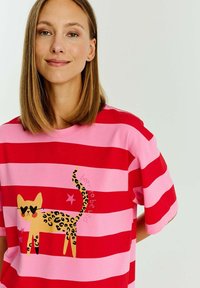 Krátký rukáv, červené a růžové pruhované tričko s grafikou žluté kočky se leopardími skvrnami a hvězdami, vyrobené z měkkého bavlněného materiálu.