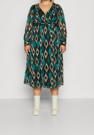 Abito lungo e modellato con una base nera e motivi geometrici verdi e beige, con scollatura avvolgente e maniche lunghe. Indossato con stivaletti bianchi alla caviglia.