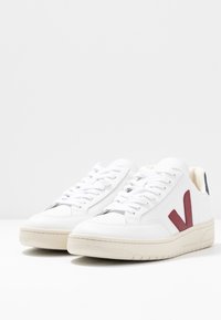 Bílé tenisky s červeným logem V, hladký kožený svršek, kulatá špička, ploché bílé tkaničky a jemná béžová gumová podrážka. Minimalistický design.