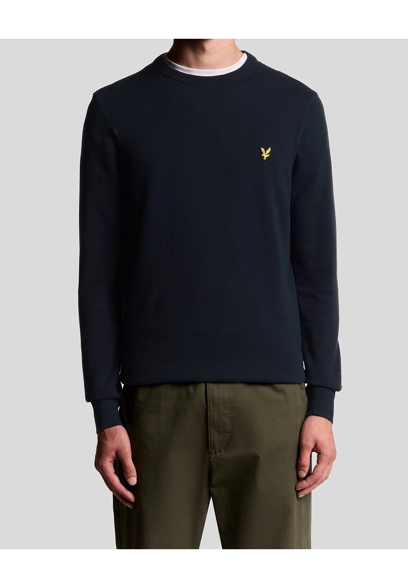 Homme portant un pull bleu marine à col rond avec un petit logo d'aigle jaune sur la poitrine et un pantalon vert olive, debout devant un fond uni.