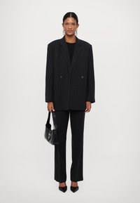 Blazer double-breasted à rayures noires avec un pantalon assorti, présentant une silhouette structurée, des revers tailleur nets et des talons noirs élégants.