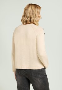 Beiger Strickpullover mit strukturierter Oberfläche, geripptem Saum und dekorativen Knöpfen an der Schulter. Kombiniert mit dunklen Jeans.