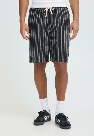 Moški, ki nosi črno-belo navpično črtaste kratke hlače z zavezovanjem, bele nogavice in črne Adidas športne copate, stoji ob preprostem ozadju.