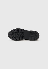 Sole de cizmă din cauciuc negru, cu crampoane adânci, având modele rectangulare și încrucișate, aflată întinsă pe un fundal deschis și simplu.