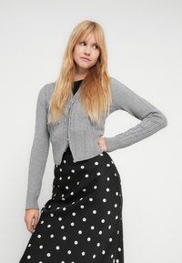 Grå strikket cardigan med teksturert forside, med lange ermer og en kort design, kombinert med et svart skjørt med polkadotter.