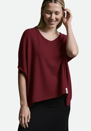 Smiling woman with blonde hair wearing a loose burgundy top and black skirt, touching her hair with one hand, relaxed posture.