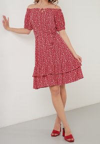 Femme portant une robe rouge à fleurs, épaules dénudées, jupe à volants, posant avec une main contre le mur et des sandales rouges à talons sur un sol gris.