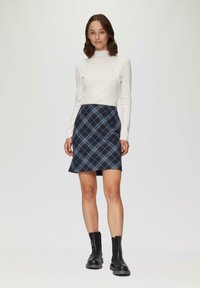 Biała, prążkowana sweterowa bluzka z długim rękawem zestawiona z granatową spódnicą w kratę z niebieskimi liniami. Modelka ma na sobie czarne botki za kostkę. Proste tło.