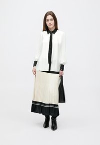 Weiche weiße Bluse mit schwarzem Kragen und Ärmeln, kombiniert mit einem plissierten beigen und schwarzen Rock, ergänzt durch eine schwarze Handtasche und kniehohe Stiefel.