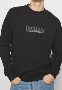Felpa nera con scollo rotondo, caratterizzata da un logo "KARL LAGERFELD" tempestato di cristalli sul petto. Tessuto morbido e polsini a costine.
