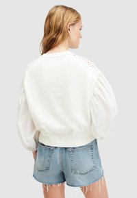 Sudadera blanca con mangas acanaladas y voluminosas y detalles recortados en los hombros, combinada con pantalones cortos de mezclilla. Tejido suave y diseño corto.
