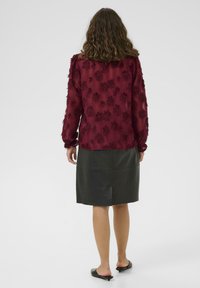 Blusa bordeaux tessuta con motivo floreale e maniche lunghe, abbinata a una gonna di pelle nera con taglio dritto e finitura liscia.