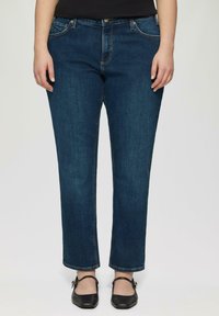 Jeans en denim de couleur bleu foncé, avec des jambes droites, un design à cinq poches, des coutures classiques, portés avec des ballerines noires.