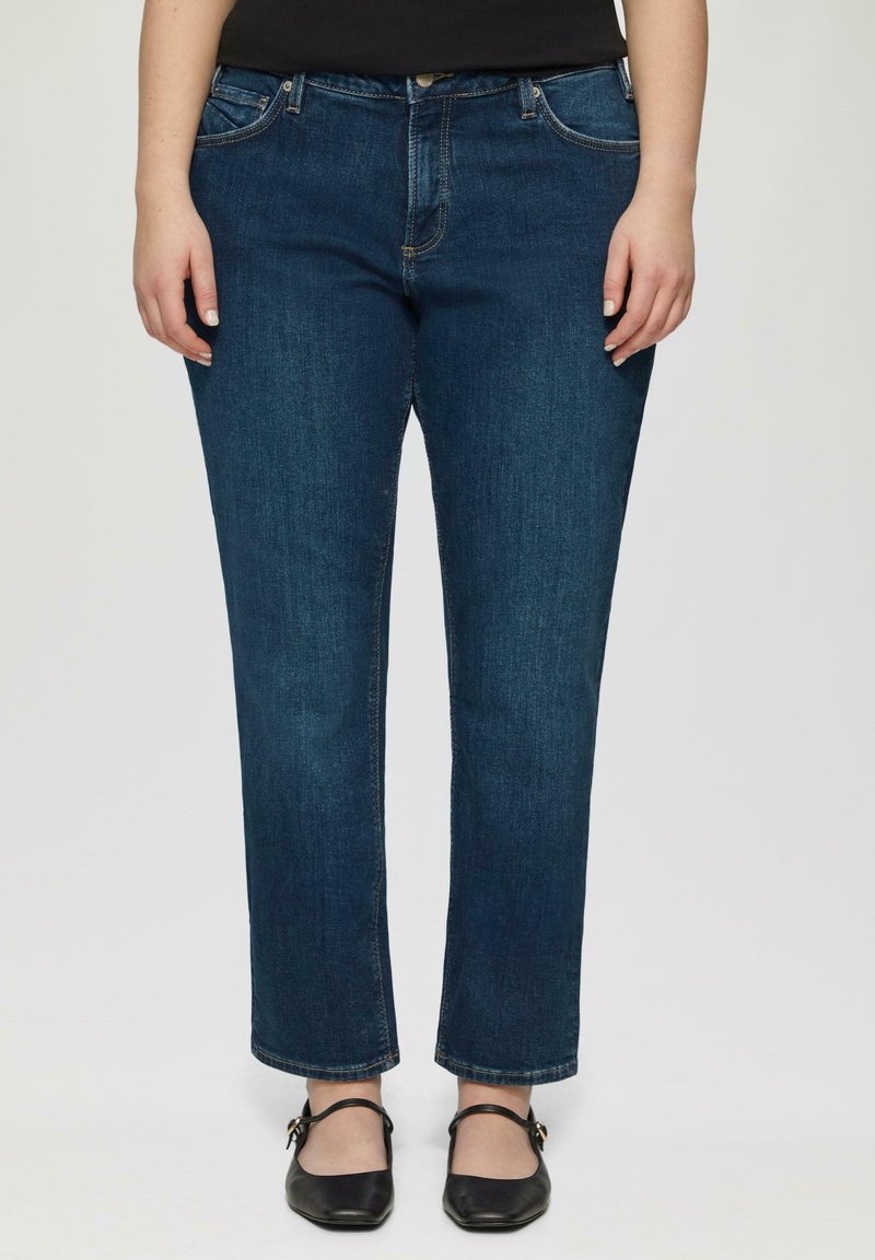 Jeans en denim de couleur bleu foncé, avec des jambes droites, un design à cinq poches, des coutures classiques, portés avec des ballerines noires.