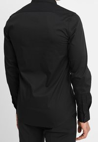 Maglietta nera a maniche lunghe realizzata in tessuto liscio, con colletto standard, polsini con bottone e orlo arrotondato. Nessun motivo o accento visibile.