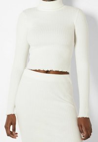 Personne à la peau foncée portant un crop top blanc à manches longues côtelé et une jupe taille haute assortie avec un détail de ourlet ondulé.