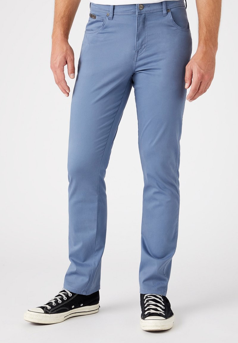 Homme portant un pantalon slim bleu clair et des baskets basses noires, debout sur un sol blanc avec un fond uni.
