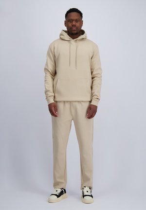 Man staat met het gezicht naar voren, draagt een beige hoodie en bijpassende joggingbroek met witte en zwarte sneakers, tegen een effen lichte achtergrond.