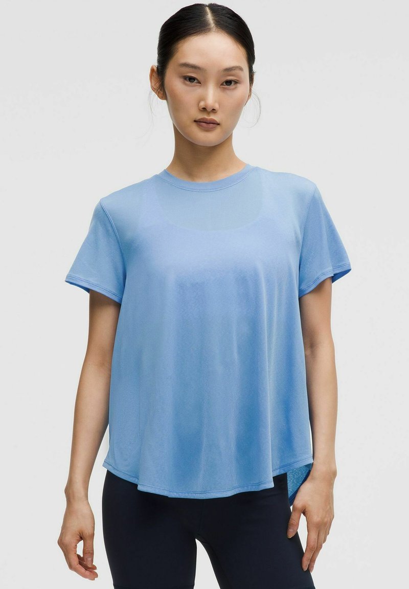 Tricou scurt cu mânecă scurtă, de culoare albastru deschis, confecționat dintr-un material neted, cu un croi lejer, decolteu rotund și o margine a spatelui mai lungă. Design simplu, fără modele.