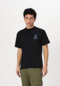 T-shirt en coton noir avec un graphique triangulaire au motif de flamme en bleu et blanc sur la zone gauche de la poitrine. Manches courtes, col rond.