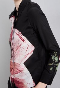 Camicia nera a maniche lunghe con una grande stampa di fiori rosa sul davanti e foglie verdi sulle maniche, indossata da una persona su uno sfondo grigio.
