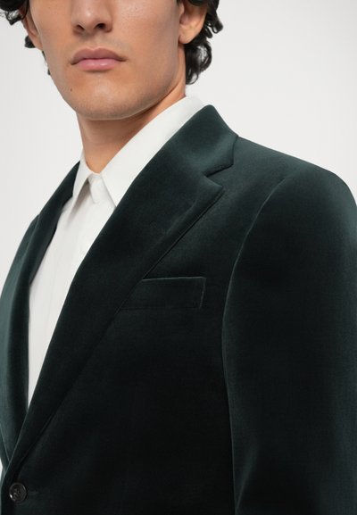 Tmavě zelený sametový blazer s klopovým límcem, zapínáním na jediný knoflík a náprsní kapsou. Pod ním bílé košili s límečkem.