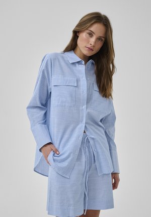 Giovane donna che indossa una camicia a righe azzurre e bianche con bottoni e pantaloncini coordinati con coulisse, posando con la mano in tasca.