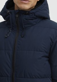 Donkerblauwe gewatteerde jas met een capuchon, voorzien van een rits aan de voorkant en trekkoordaccenten. Glad textuur met gestikte secties voor isolatie.