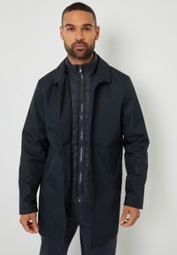 Sort vandtæt frakke med krave, der har et lynlåset indre pufferskef. Glat tekstur, sidelommer og en moderne silhuet.