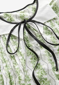 Blusa bianca con motivo floreale verde, dotata di pieghe, colletto nero e fiocco. Tessuto testurizzato con dettagli neri lungo i bordi.