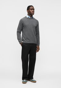 Homme debout portant un pull gris par-dessus une chemise col bleu clair, un pantalon noir et des baskets teal avec des semelles en caoutchouc, sur un fond uni.