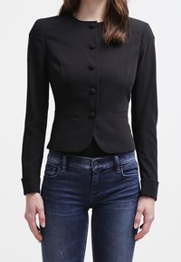 Svart skräddarsydd jacka med rund krage, sex framknappar och långa ärmar. Kombinerad med mörkblå jeans ser tyget slätt och strukturerat ut.