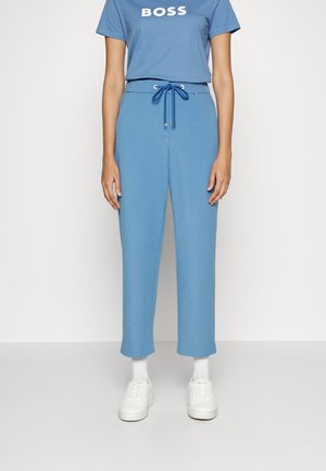 Personne portant un pantalon bleu à cordon, un t-shirt bleu avec le texte "BOSS", des chaussettes blanches et des baskets blanches, debout sur un fond uni.