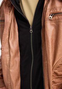 Chaqueta de cuero marrón con una capa interior negra, que presenta herrajes de cremallera plateados y detalles de costura en el lado derecho.