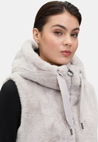 Jasnoszary wdzianko z futra syntetycznego z dużym kapturem, posiadające srebrne detale i regulowane szare sznureczki. Miękka tekstura i bezrękawnikowy krój.