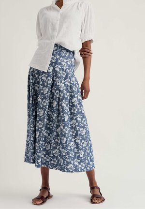 Žena oblečená v bielej textúrovanej blúzke na gombíky s nafúknutými rukávmi, modrej midi sukni s kvetinovým vzorom a hnedých sandáloch s remienkom okolo členka, stojaca na jednoduchom pozadí.